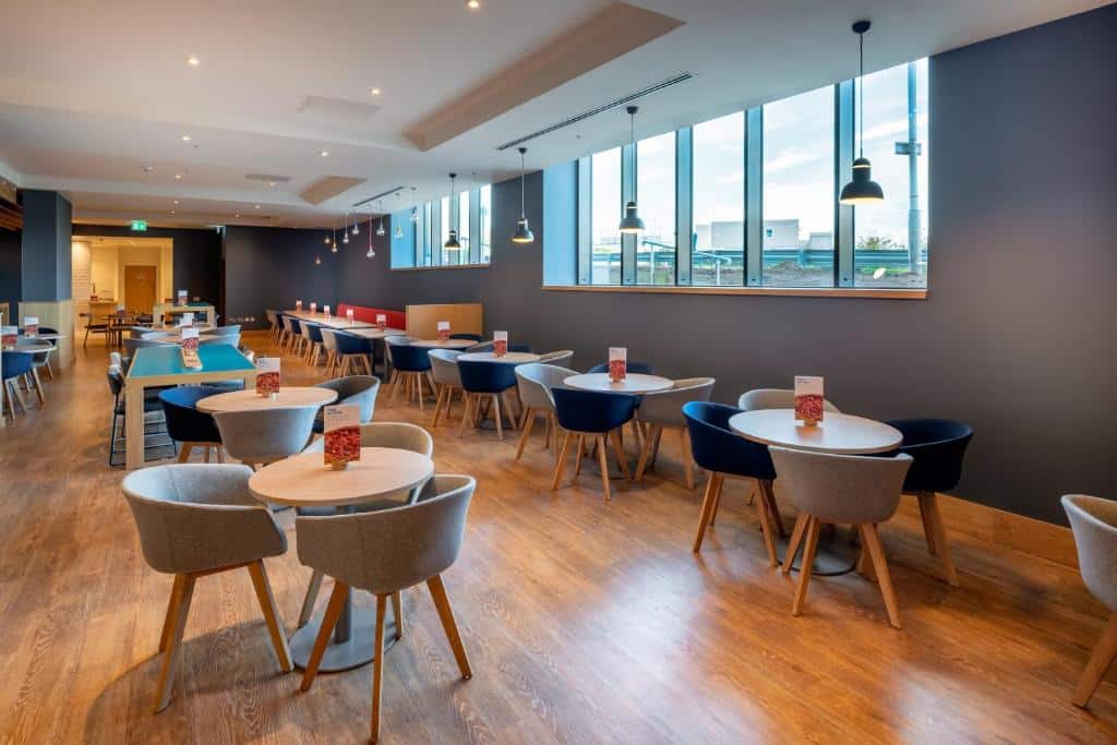Salão de refeições do Holiday Inn Express - London Heathrow T4, an IHG Hotel com pequenas mesas redondas, cadeiras estofadas, algumas janelas e lustres