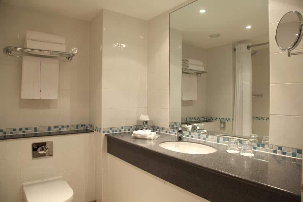 Banheiro do Holiday Inn London Bloomsbury, an IHG Hotel com prateleiras com toalhas brancas, um espelho, uma pia ampla e um vaso sanitário, o box não é de vidro, para representar hotéis Mercure em Londres