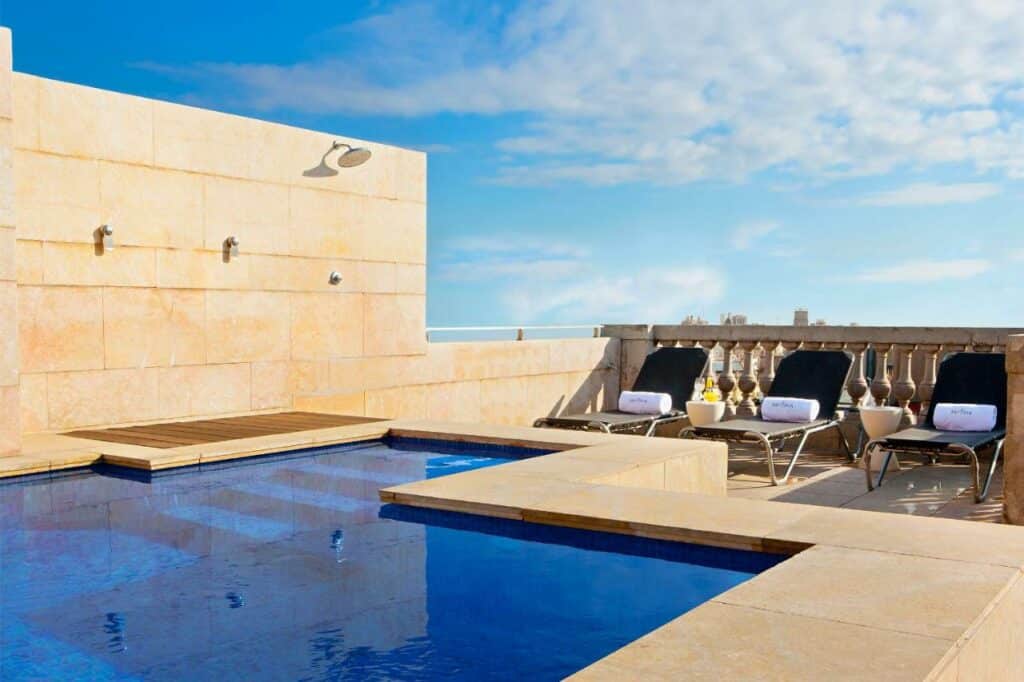 Área da piscina do Hotel Astoria, uma das recomendações de hotéis baratos em Barcelona. Três espreguiçadeiras pretas estão encostadas na balaustrada do outro lado da piscina e têm toalhas brancas dobradas em cima. Há uma ducha ao lado da piscina. A foto foi tirada durante o dia e o céu está azul.