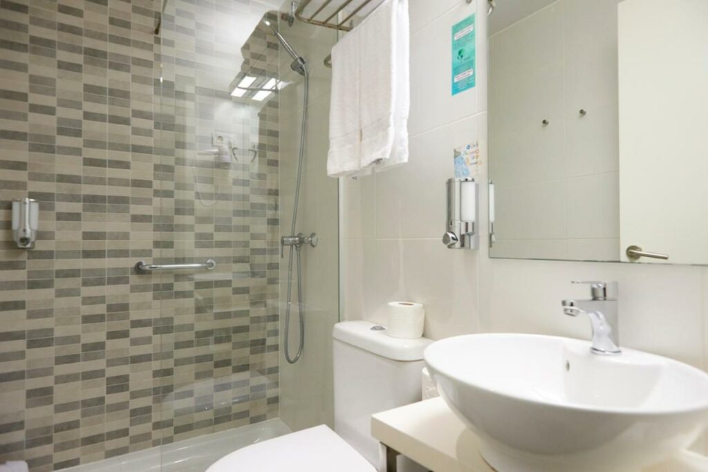 Banheiro do Hotel BESTPRICE Diagonal. A área do chuveiro é protegida por um box de vidro e tem uma barra de apoio. A privada está entre o box e a pia, que tem um espelho na parede acima. Há um suporte de toalhas em cima da privada.
