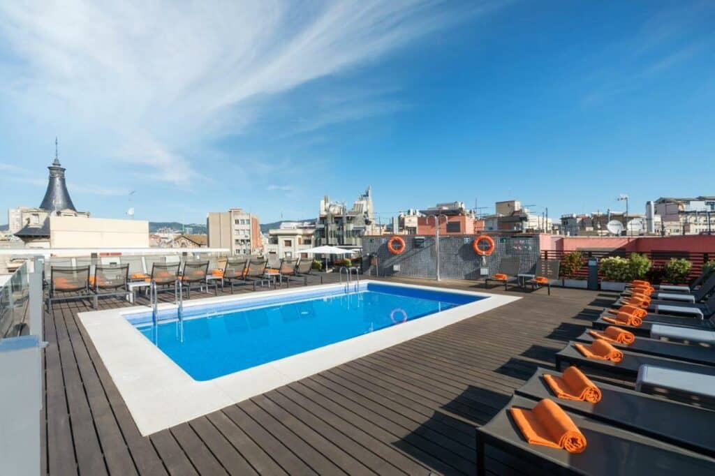 Piscina do Hotel Jazz, um dos melhores hotéis em Barcelona. Espreguiçadeiras tem toalhas dobradas ao longo da piscina, e o terraço tem vista para a cidade.