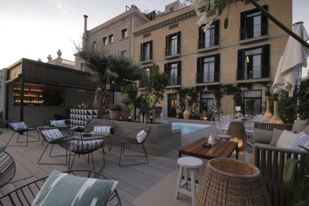 Área da piscina do Hotel Oasis, um dos melhores hotéis em Barcelona. Há mesinhas com cadeiras e sofás ao redor, e a piscina está ao longe, perto do prédio com janelas.