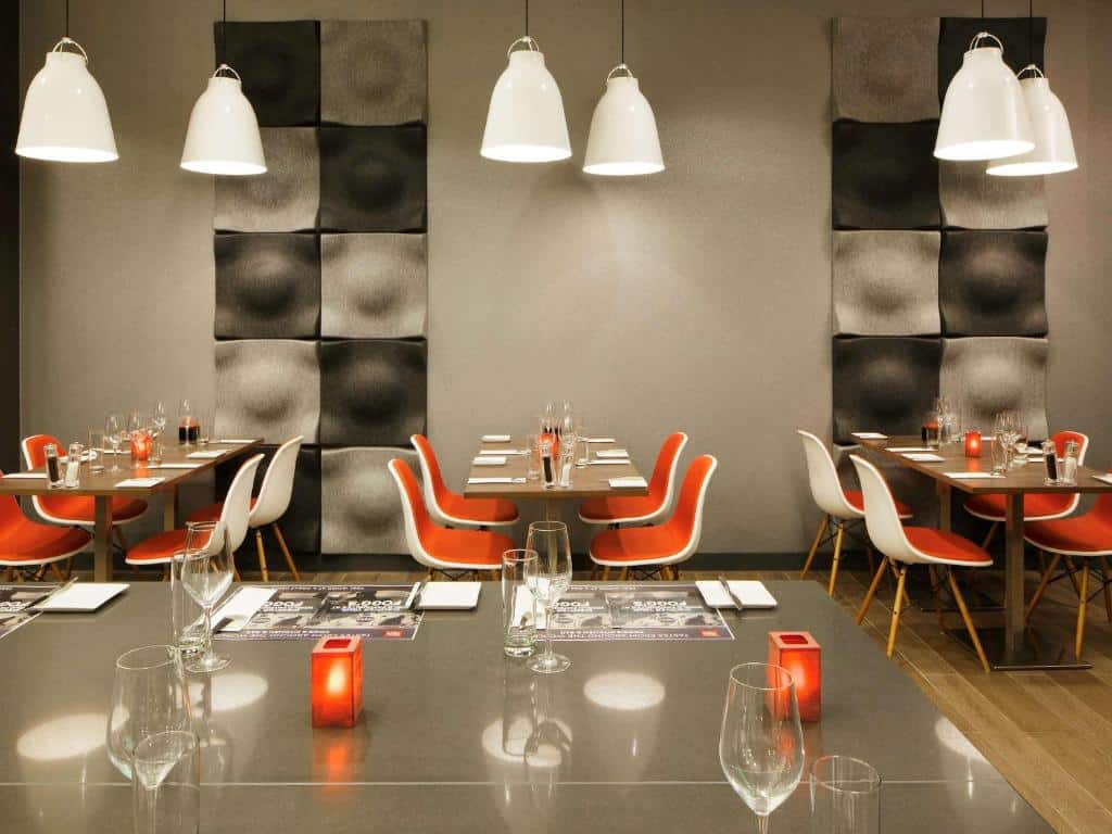 Salão de refeições do ibis London Blackfriars com mesas de madeira com quatro cadeiras vermelhas, chão de madeira e alguns lustres brancos