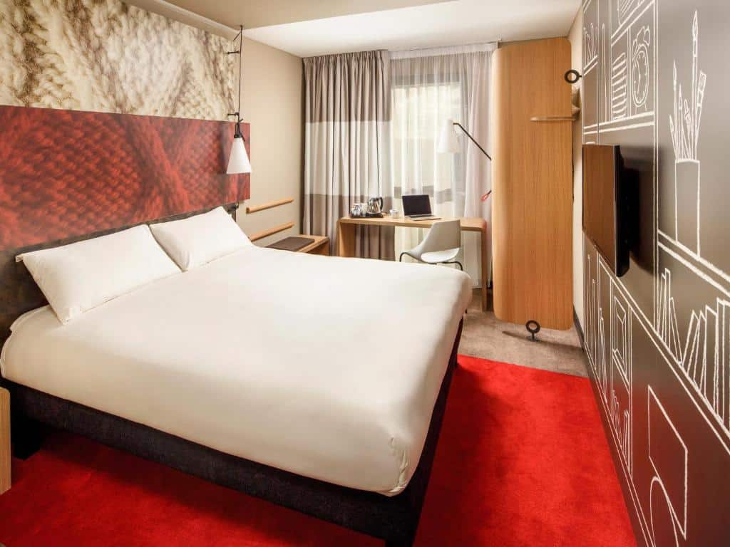 Quarto do ibis London Canning Town com uma cama de casal, uma janela com cortinas, uma bancada de madeira com uma cadeira giratória, um tapete vermelho e uma televisão, para representar hotéis Ibis em Londres