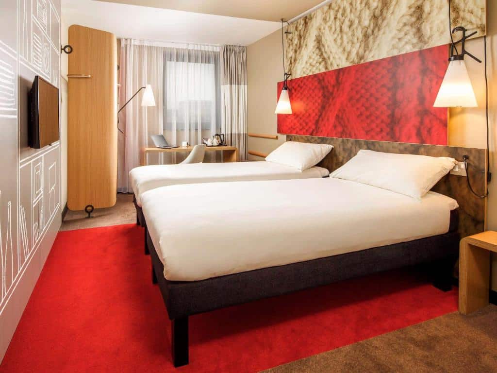 Quarto do ibis London Docklands Canary Wharf com duas camas de solteiro, uma janela com cortinas, uma mesa de escritório com uma cadeira, um tapete vermelho e uma televisão