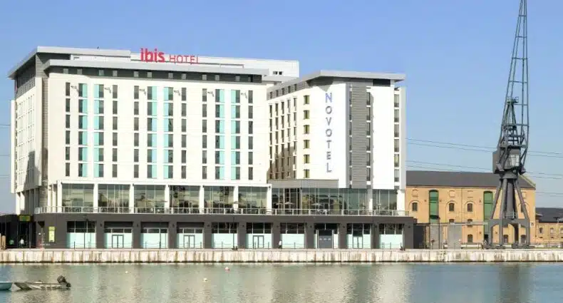O ibis London Excel-Docklands na beira do rio, com quatorze andares e o nome em destaque em vermelho, para representar hotéis Ibis em Londres