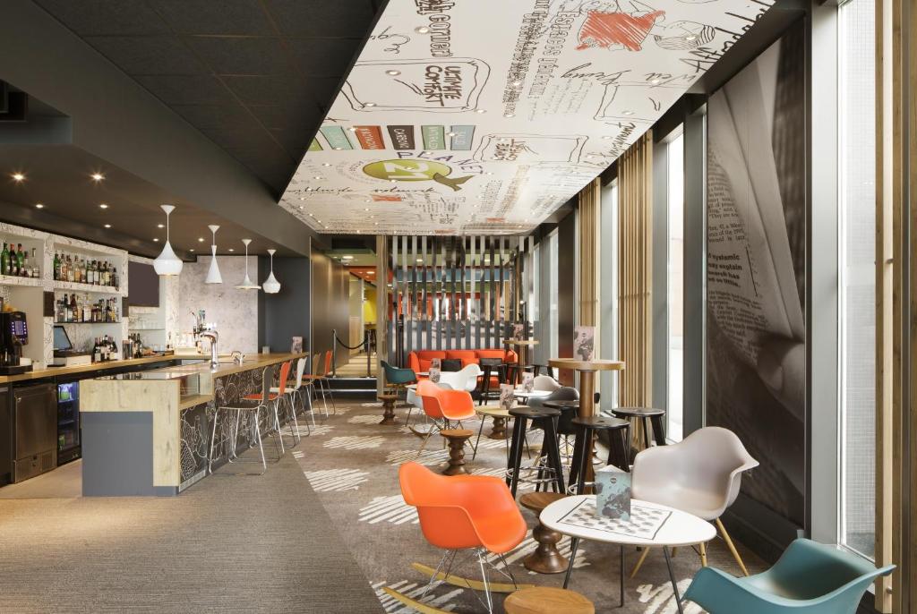 Salão de refeições do ibis London Excel-Docklands com mesas redondas, poltronas coloridas, uma bancada em estilo de bar com banquetas, ambiente decorado e despojado