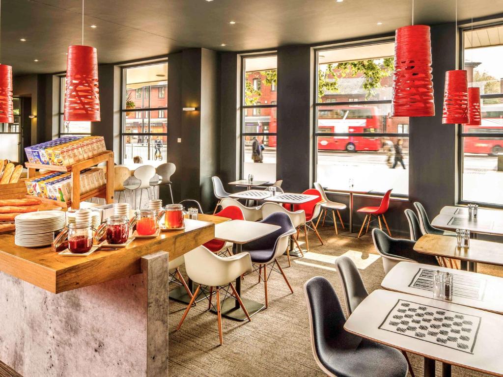 Salão de refeições do ibis London Stratford com mesas brancas, cadeiras coloridas, lustres vermelhos e uma bancada de madeira com itens de café da manhã