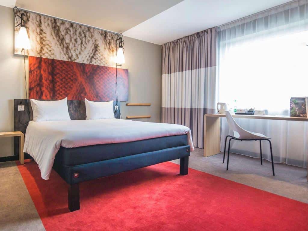 Quarto espaçoso do ibis London Stratford com uma cama de casal, uma janela com cortinas, um tapete vermelho, uma bancada de trabalho e uma cadeira, para representar hotéis Ibis em Londres