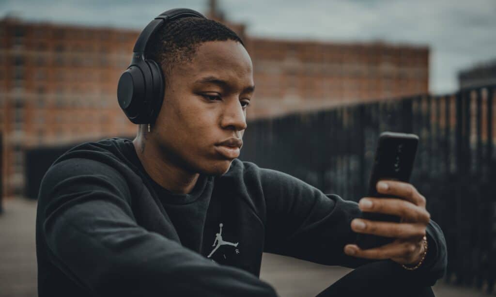 rapaz negro sentado, usando um headphone preto, camiseta preta e segurando um celular android preto com a mão esquerda. Ele está olhando para a tela do celular. Atrás dele é possível enxergar algumas construções antigas