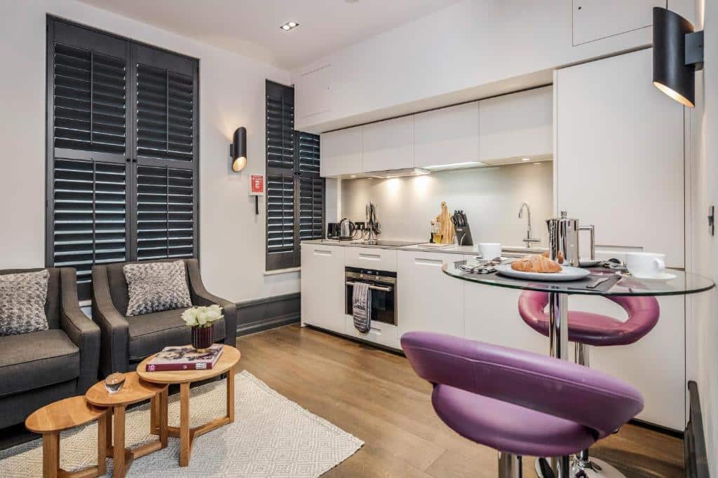 Cozinha do Kula London - Covent Garden Garrick St com duas janelas, móveis brancos, duas poltronas, uma mesinha redonda com duas cadeiras roxas e o chão é de madeira