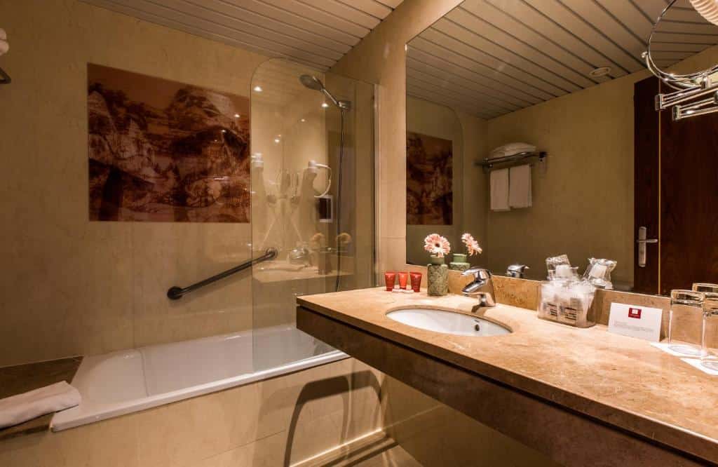Banheiro do Leonardo Hotel Barcelona Gran Via. Uma banheira com barra de apoio e chuveiro está protegida por um box de vidro. Há um quadro na parde acima da banheira. A pia de mármore tem amenidades de banho, copos de vidro e um grande espelho na parede acima.