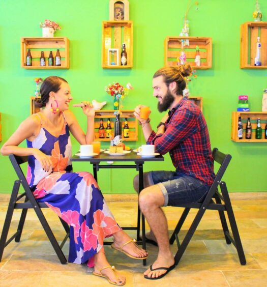 Um casal sentado em uma mesa de madeira em um ambiente compartilhado do Local Hostel Manaus tomando café da manhã em um ambiente todo decorado, para representar o Local Hostal Manaus