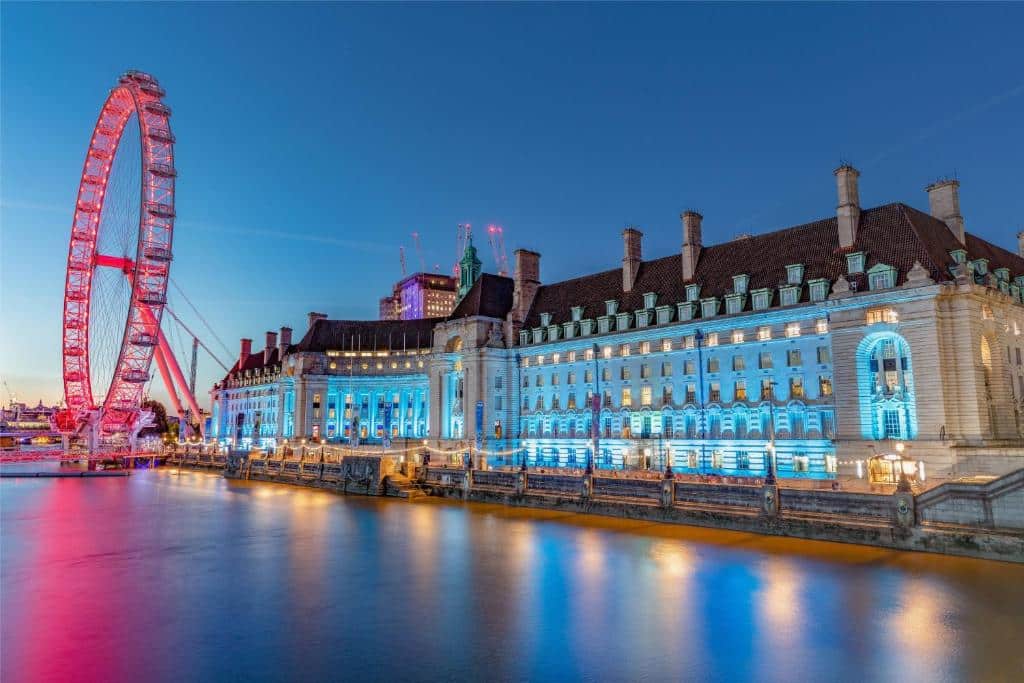 Prédio do London Marriott Hotel County Hall inteiro iluminado com luzes azuis, de frente para o rio Tâmisa e, do lado esquerdo, há a London Eye iluminada com luzes rosas