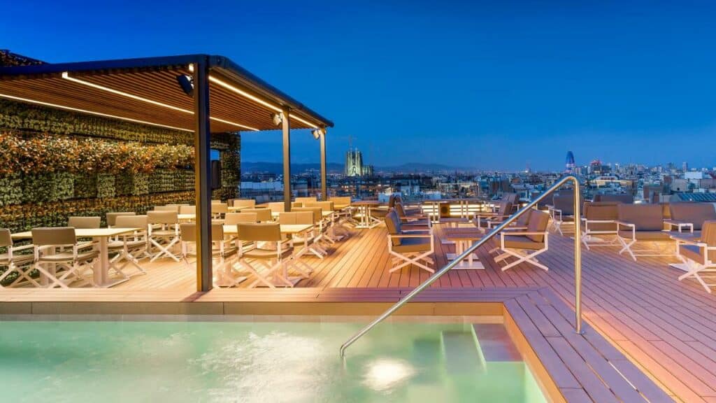 Piscina do Majestic Hotel & Spa Barcelona GL, uma das recomendações de hotéis românticos em Barcelona. Uma barra de apoio leva aos degraus e à água. Ao fundo, mesas e cadeiras estão ao redor no deque. Há vista para a cidade ao longe.