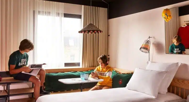 Quarto no Mama Shelter London - Shoreditch com uma janela com cortinas, uma cama, um sofá de canto e dois meninos sentados no ambiente, para representar hotéis para família em Londres