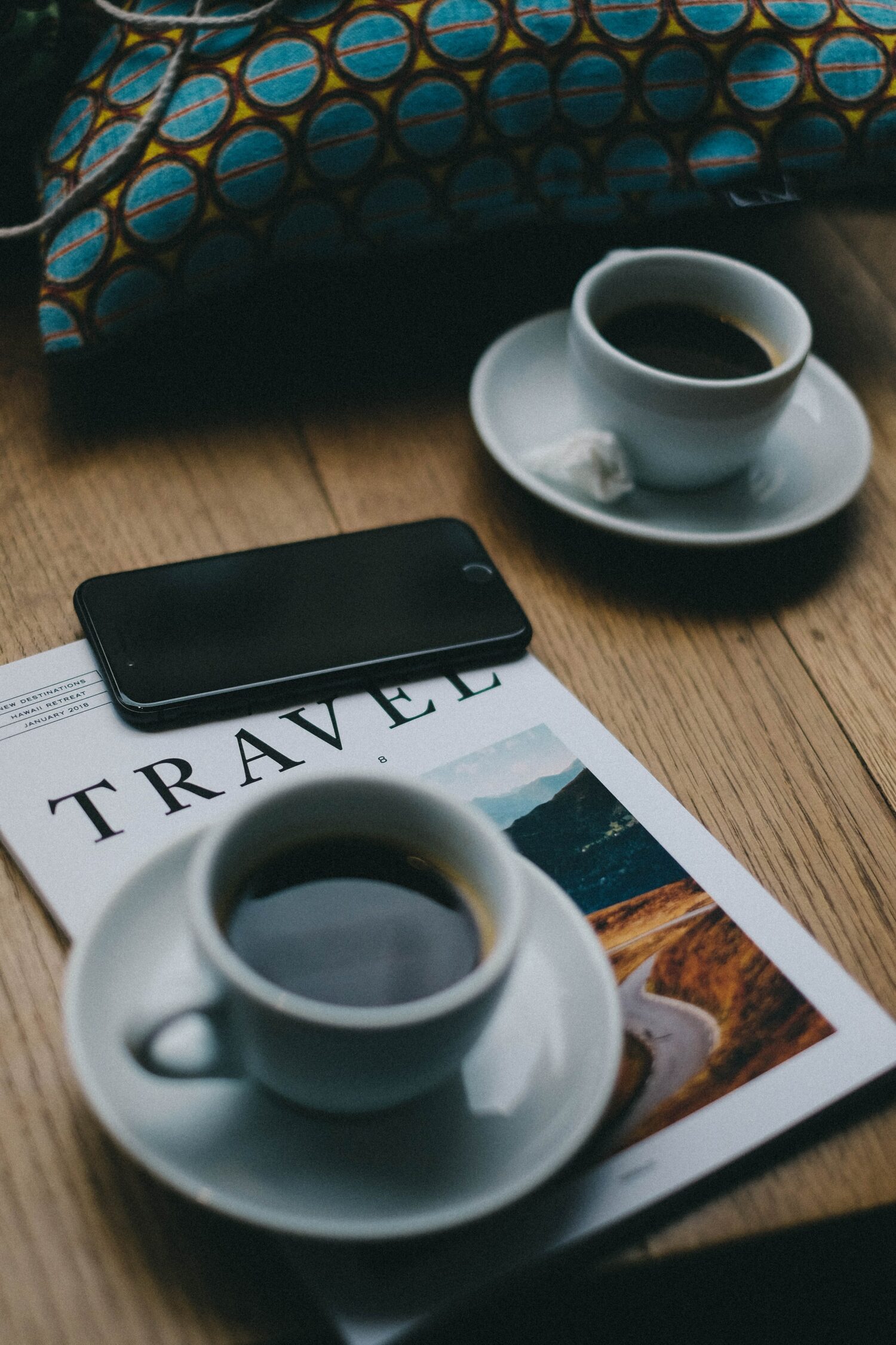 mesa de madeira com um celular android preto em cima de uma revista de viagem com duas xícaras brancas com pírex e café dentro. É usada para ilustrar a importância de contratar um chip de celular para viagem internacional
