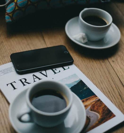 mesa de madeira com um celular android preto em cima de uma revista de viagem com duas xícaras brancas com pírex e café dentro. É usada para ilustrar a importância de contratar um chip de celular para viagem internacional