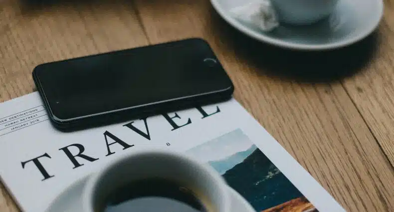 mesa de madeira com um celular android preto em cima de uma revista de viagem com duas xícaras brancas com pírex e café dentro. É usada para ilustrar a importância de contratar um chip de celular para viagem internacional