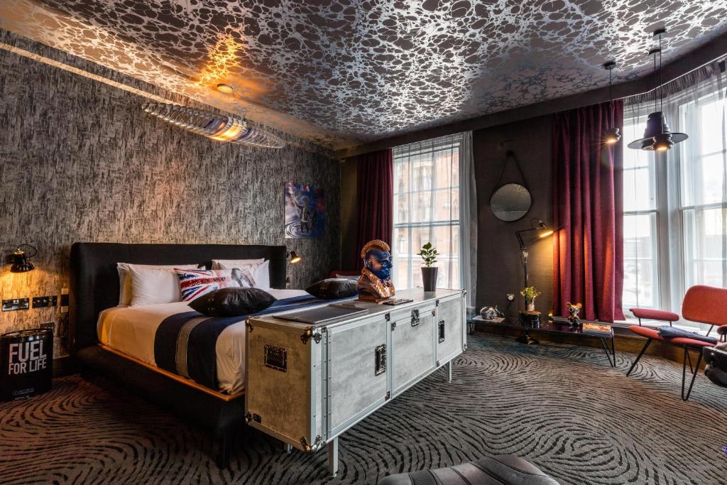 Quarto amplo do Megaro Hotel com uma cama de casal, duas janelas com cortinas, carpete no chão, um móvel que se parece com um baú e alguns itens de decoração, para representar hotéis bem localizados em Londres