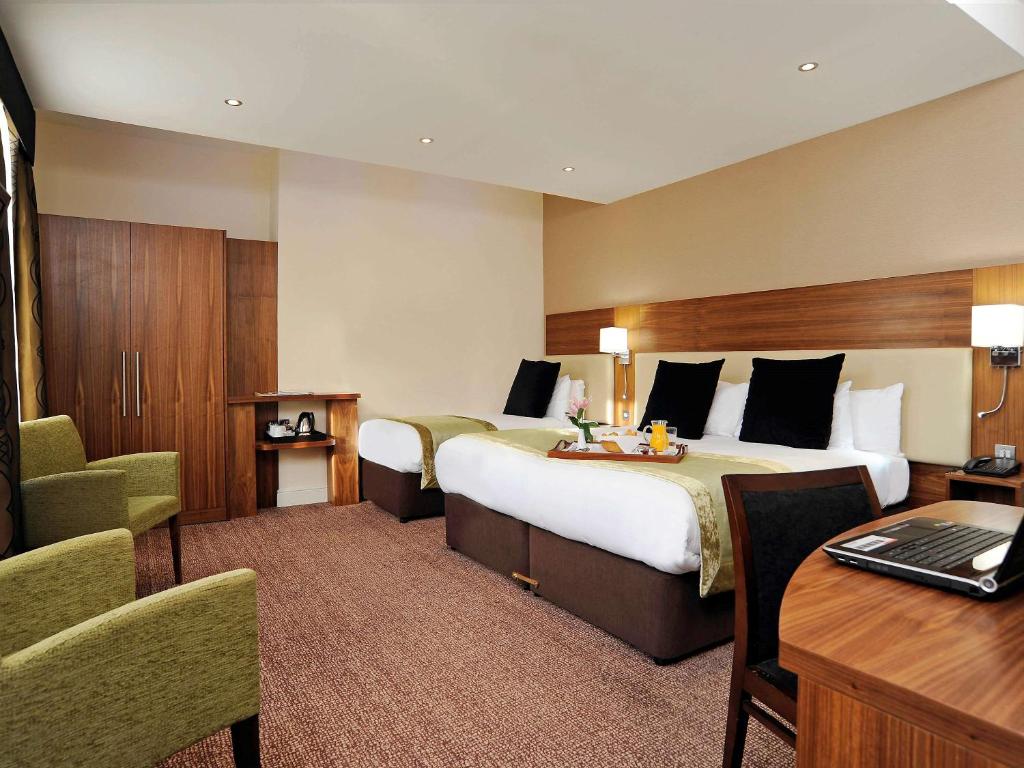 Quarto do Mercure London Bloomsbury Hotel com uma cama de casal, uma de solteiro, duas poltronas, um pequeno armário, carpete e uma mesa de escritório