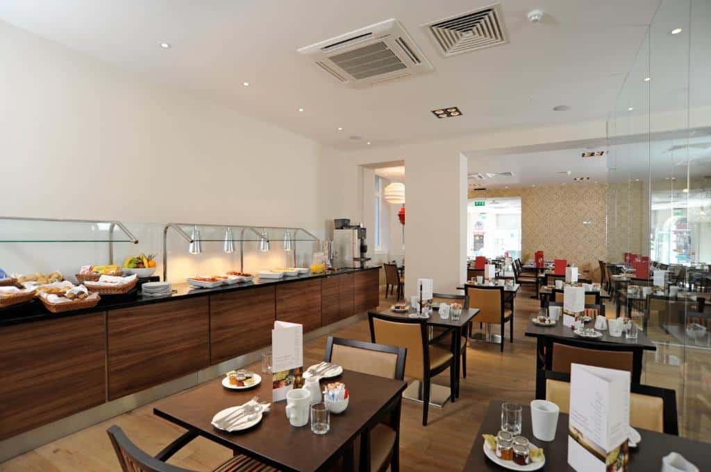 Sala de refeições do Mercure London Bloomsbury Hotel com mesas quadradas de madeira, cadeiras, e do lado há um balcão de buffet, para representar hotéis Mercure em Londres