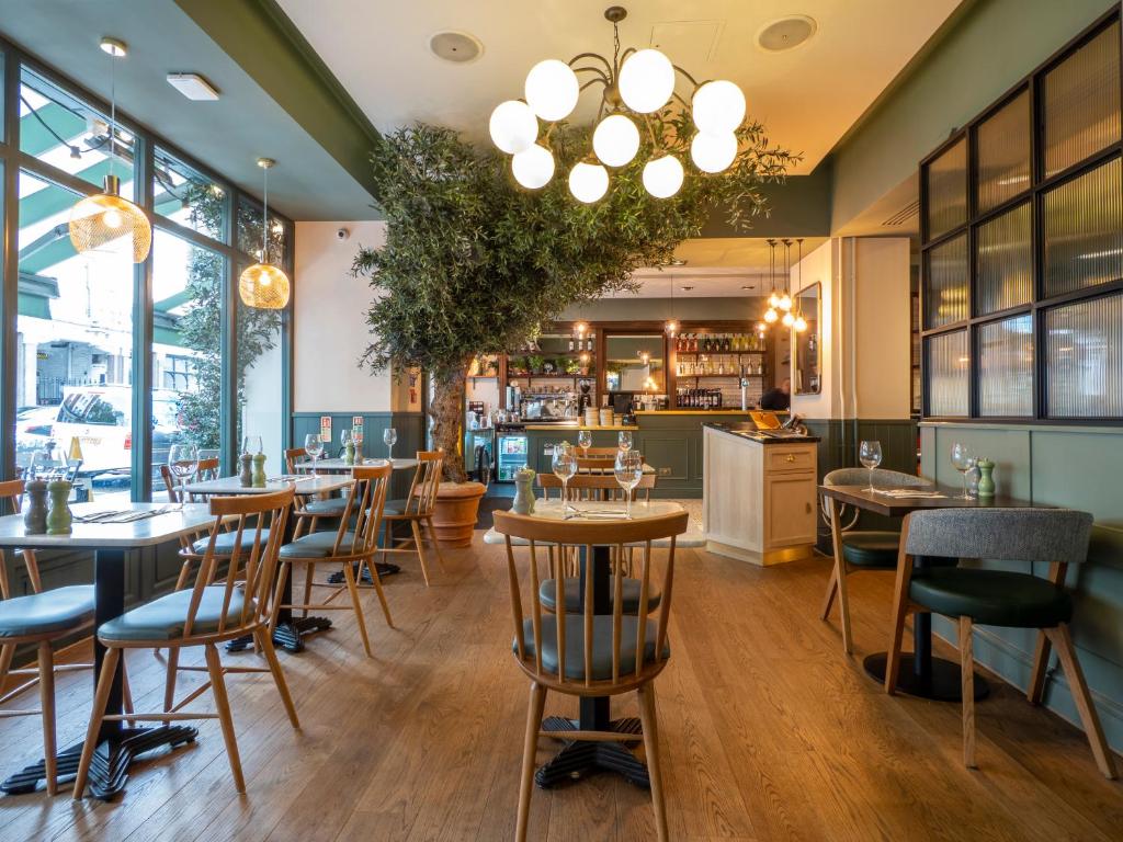 Sala de refeições do Mercure London Hyde Park Hotel com mesas de madeira, cadeiras de madeira, uma árvore, um lustre com lâmpadas redondas e uma janela com vista para a rua, para representar hotéis Mercure em Londres