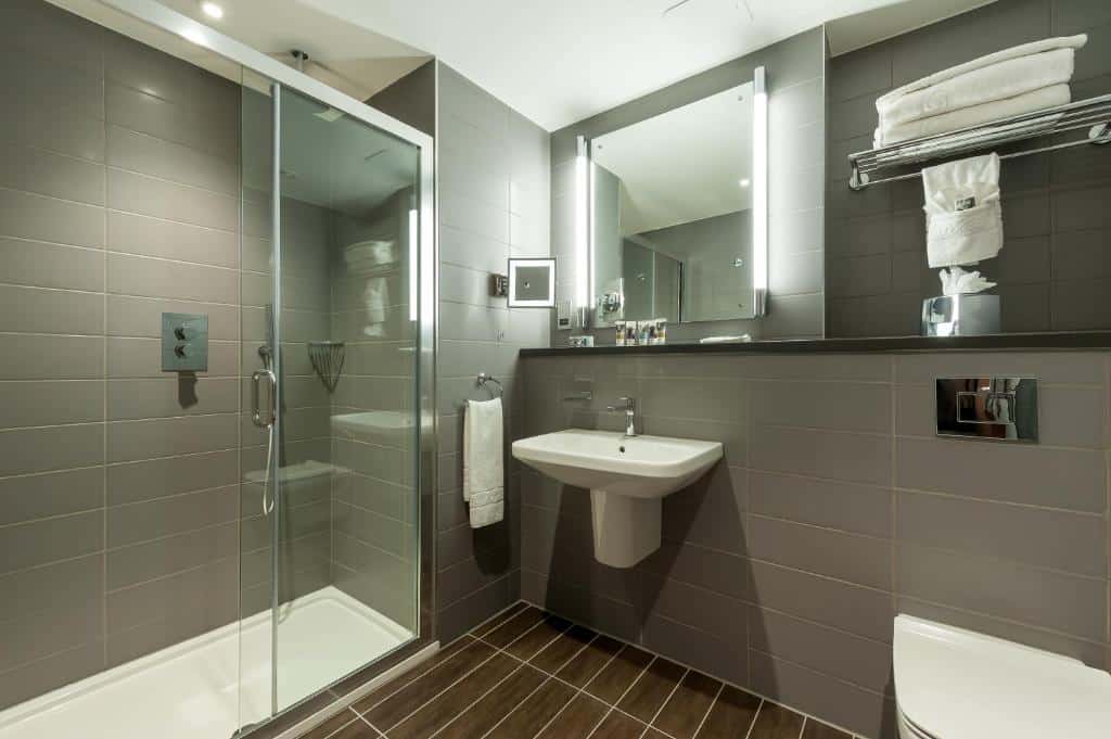 Banheiro do Mercure London Paddington Hotel com um box de vidro, uma pia sem móvel, um espelho e uma prateleira com toalhas brancas, para representar hotéis Mercure em Londres