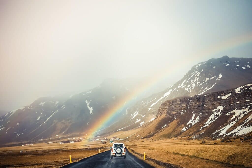 Estrada na Islândia, com carro parado nela, vegetação de tom amarronzado em ambos os lados, montanhas ao fundo com pontos de neve, e um arco-íris sobre o carro