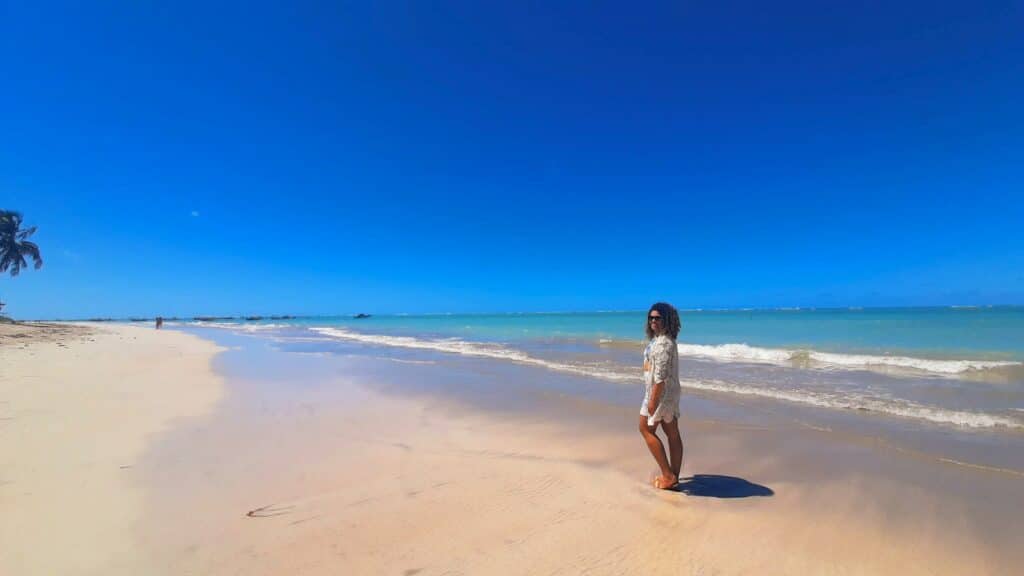 Mulher, com o corpo de lado olhando para frente, em uma praia de São Miguel dos Milagres com areia clara e mar azul ao fundo