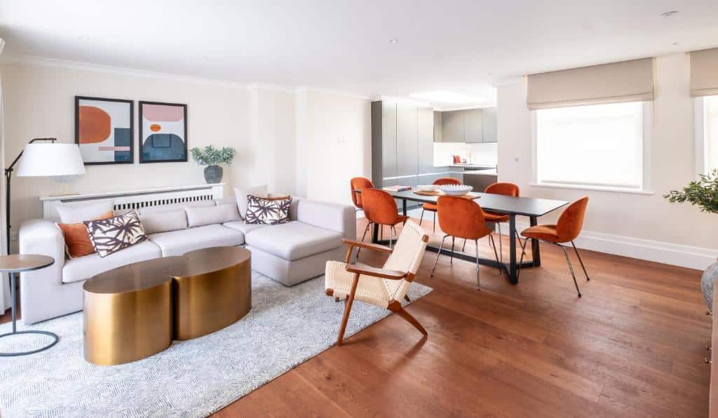 Sala de estar do Native Mayfair com chão de madeira, mesa com seis lugares, janelas com persiana, uma sofá com almofadas coloridas e um tapete cinza
