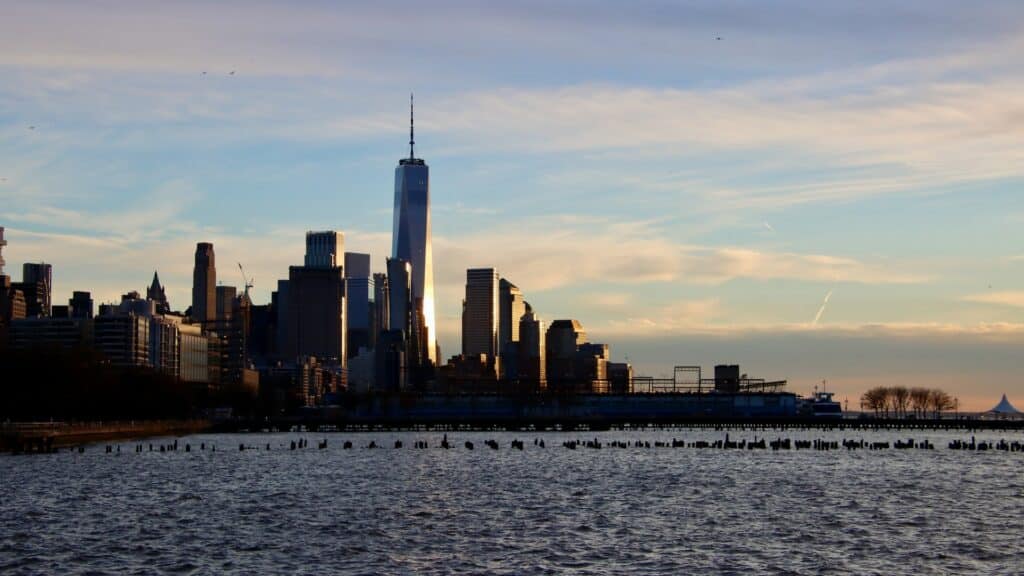 Horizonte de Nova York, com diversos prédios e arranha-céus ao fundo, e destaque para o One World Trade Center, e água do rio Hudson à frente, para ilustrar o post sobre como tirar visto americano