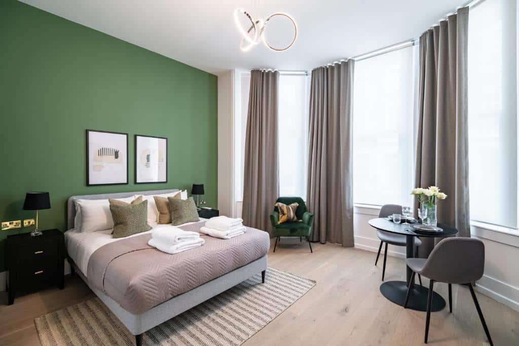 Quarto do No. 2 Queensberry Place by Stayo com uma cama de casal, três janelas amplas com cortinas, um tapete, uma pequena mesa com duas cadeiras, e alguns itens de decoração como quadros e um lustre, para representar airbnb em Londres