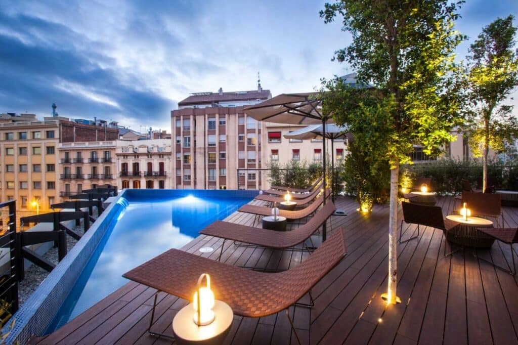 Cobertura do Ocean Drive Barcelona, uma das recomendações de hotéis de luxo em Barcelona. Uma pequena piscina na beira do terraço tem espreguiçadeiras intercaladas com mesinhas e guarda-sóis ao redor. Há algumas árvores e mesas com cadeiras no local. Ao fundo há vista para a cidade.