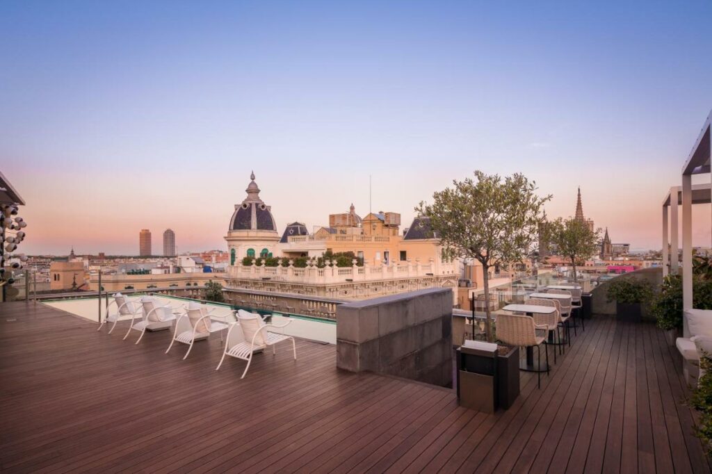 Terraço do Ohla Barcelona, um dos melhores hotéis em Barcelona. Há várias mesas com cadeiras do lado direito, e uma pequena árvore em um vaso. Do lado esquerdo, espreguiçadeiras brancas encaram uma piscina. Há uma balaustrada ao redor da cobertura, e é possível ver outros prédios da beirada.