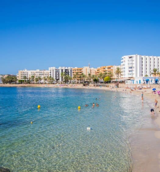 Orla da Playa De Santa Eulalia em Ibiza com o mar água claro, alguns prédios e pessoas andando, para representar praias em Ibiza