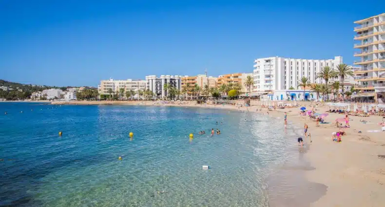 Orla da Playa De Santa Eulalia em Ibiza com o mar água claro, alguns prédios e pessoas andando, para representar praias em Ibiza
