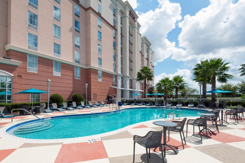 piscina com bordas arredondadas com cadeiras ao redor e a fachada atrás do hotel em tons de bege do Hampton Inn & Suites Orlando Airport at Gateway Village, um dos hotéis perto do aeroporto de Orlando