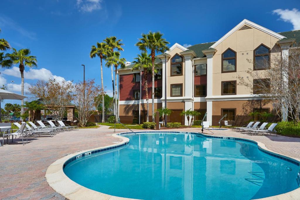 piscina com bordas arredondadas do Staybridge Suites Orlando South, um dos hotéis perto do aeroporto de Orlando, com algumas palmeiras e várias cadeiras ao redor, com a fachada do hotel ao fundo parecendo casinhas clássicas grandes