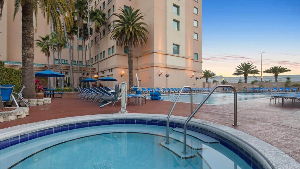 piscinas do The Florida Hotel & Conference Center in the Florida Mall, um dos hotéis para brasileiros em Orlando, sendo uma maior retangular ao fundo com cadeiras em volta e uma menor redonda de hidro, há algumas palmeiras baixas e o hotel está atrás