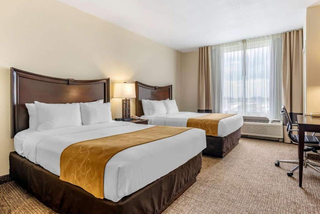 quarto do Comfort Suites Maingate East, um dos melhores hotéis em Orlando, com janela grande que pega quase toda parede com cortinas claras e um sofá abaixo, há duas camas de casal grandes com detalhes em dourado e de ambos os lados de ambas as camas há uma mesinha com luminária