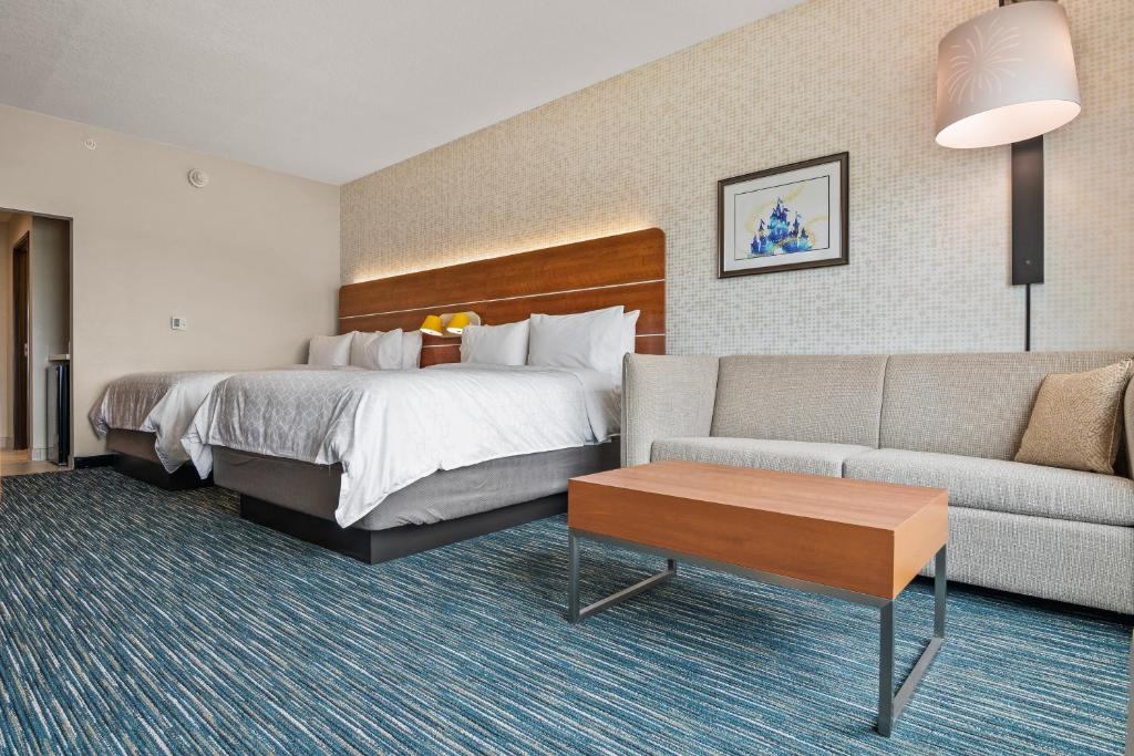 quarto amplo do Holiday Inn Express & Suites Orlando, um dos hotéis em Orlando, com duas camas de casal com luzes na cabeceira e um sofá comprido com mesinha de centro