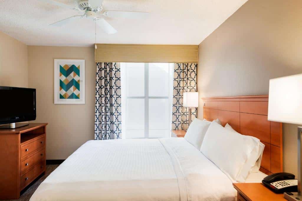 quarto do Homewood Suites by Hilton Orlando-Intl Drive/Convention Ctr com cama de casal, mesinha e luminária do lado, janela com cortina colorida e cômoda e tv à frente