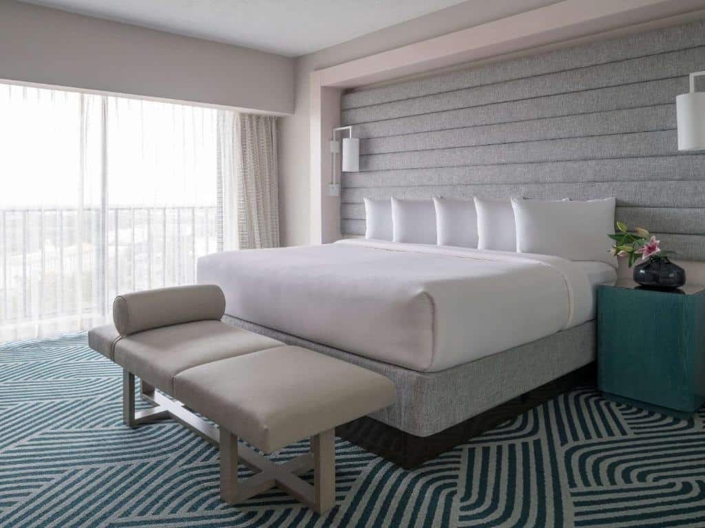 quarto do Hyatt Regency Grand Cypress Resort com carpete em detalhes azuis e cama de casal bem grande em branco, com luminárias em ambos os lados, uma mesinha e janela de vidro com varandinha e cortinas bem claras