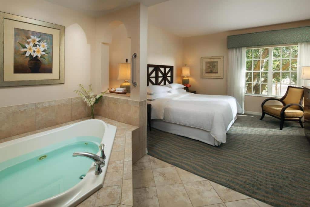 quarto com banheira do Sheraton Vistana Resort Villas, Lake Buena Vista Orlando, a banheira está em um ambiente que se comunica com o quarto de cama de casal, mesinha de luminária de ambos os lado, poltrona e janelas de vidro