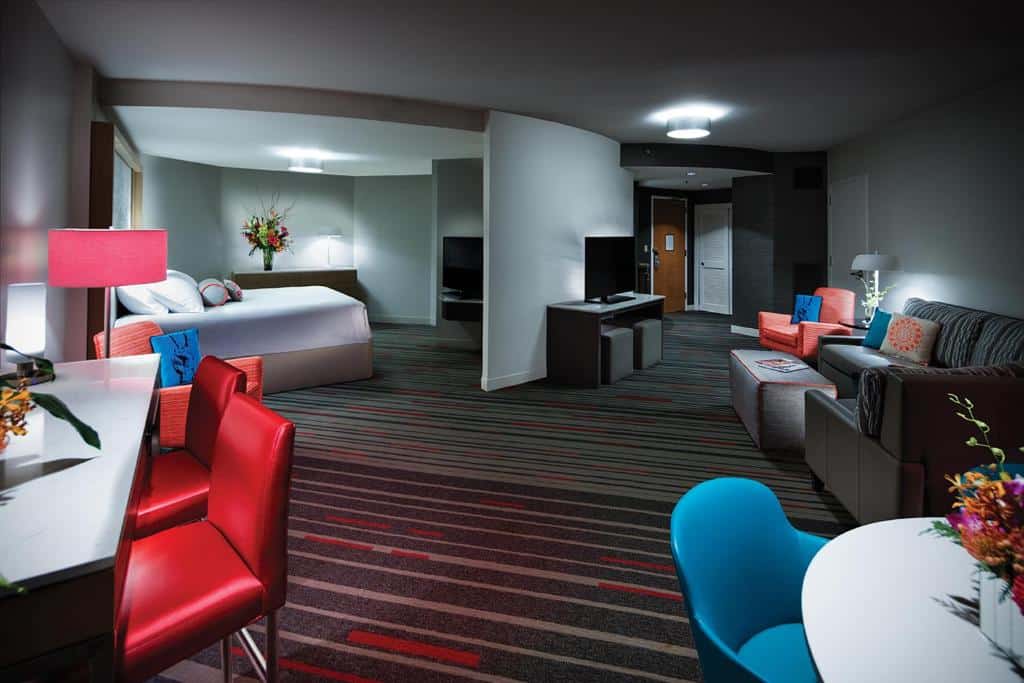 quarto grande do Universal's Hard Rock Hotel com carpete e móveis coloridos, sofá com tv na frente, mesa com cadeiras, balcão e, em um ambiente separado, uma cama de casal grande com tv na frente