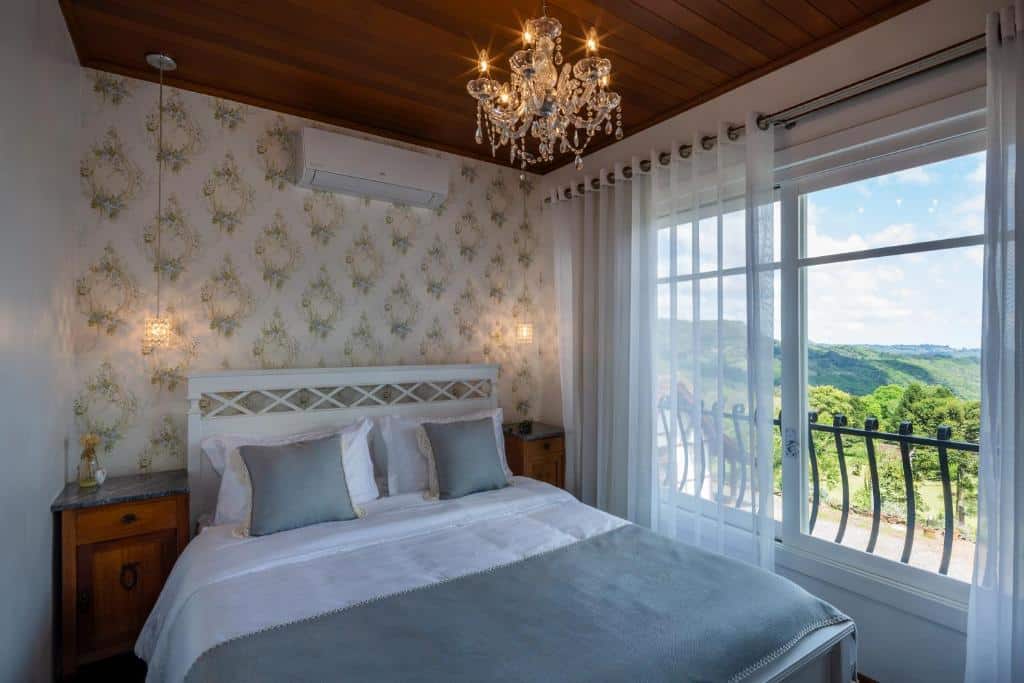 suíte da Pousada La Vista com uma cama de sala, uma parede coberta em papel de parede florido e uma janela ampla à direita com vista para a serra e um dia ensolarado.