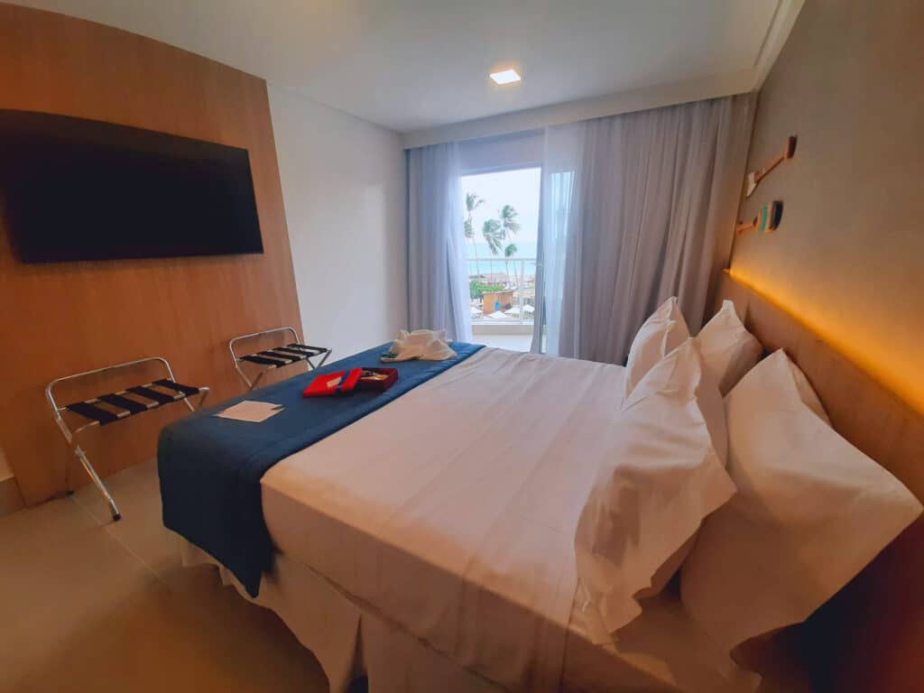 Quarto do hotel Brisa Maragogi, com uma cama de casal, TV e à direita tem uma porta de vidro com a cortina semi aberta e vista do mar e piscinas.