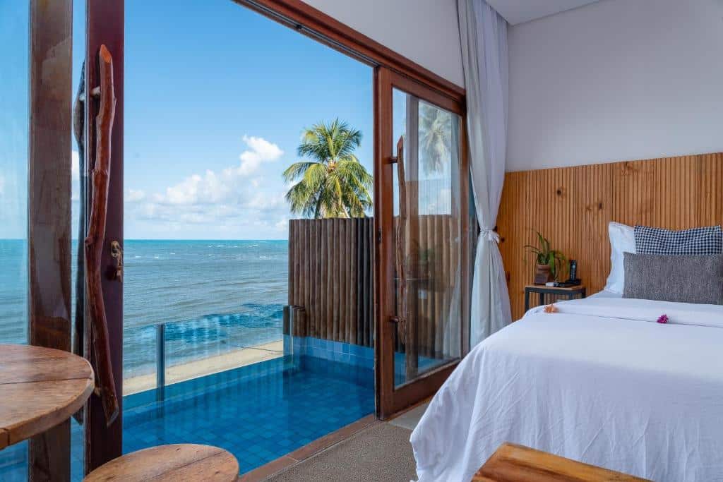 Quarto com cama king-size do Villa Pantai Boutique Hotel Maragogi, sendo que na imagem aparece metade da cama e à esquerda tem uma piscina na sacada com borda de vidro e vista do mar