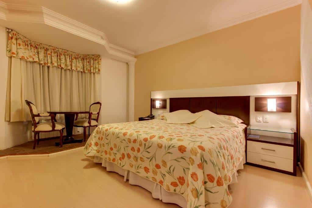 quarto simples do Hotel Glamour da Serra em Gramado com uma cama de casal e uma pequena mesa redonda de madeira com duas cadeiras estofadas e brancas, um de frente para outra.